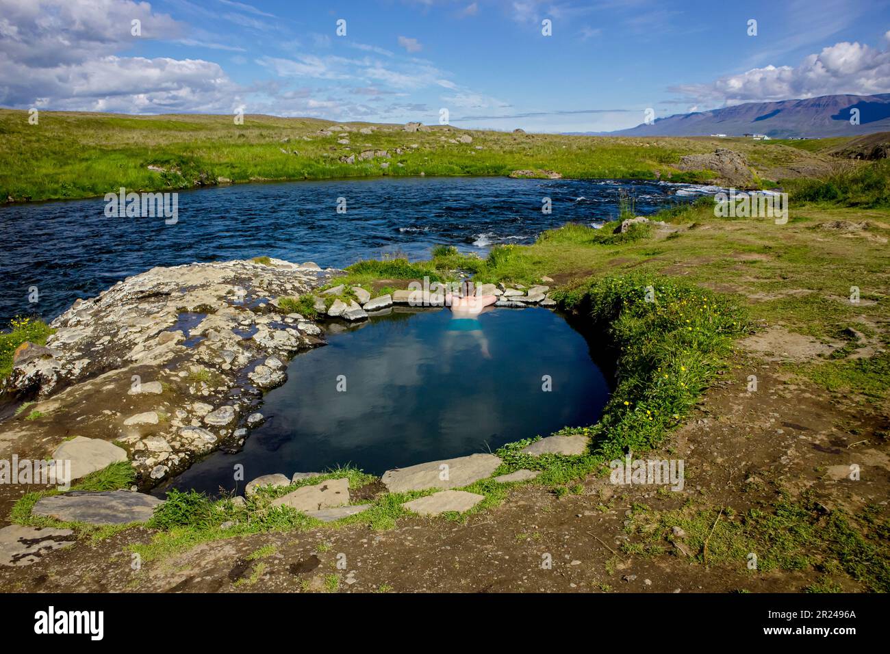 Uomo che si rilassa e fa il bagno nella piscina calda geotermica naturale riscaldata, sorgente termale calda in Islanda in estate. Verdi colline ondulate e cielo blu sullo sfondo. Foto Stock