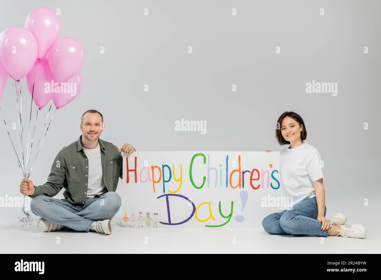 Sorridente famiglia adulta in abiti casual guardando la macchina fotografica mentre tiene palloncini e cartellone rosa festivo con felice giorno dei bambini scritta sul grigio b Foto Stock