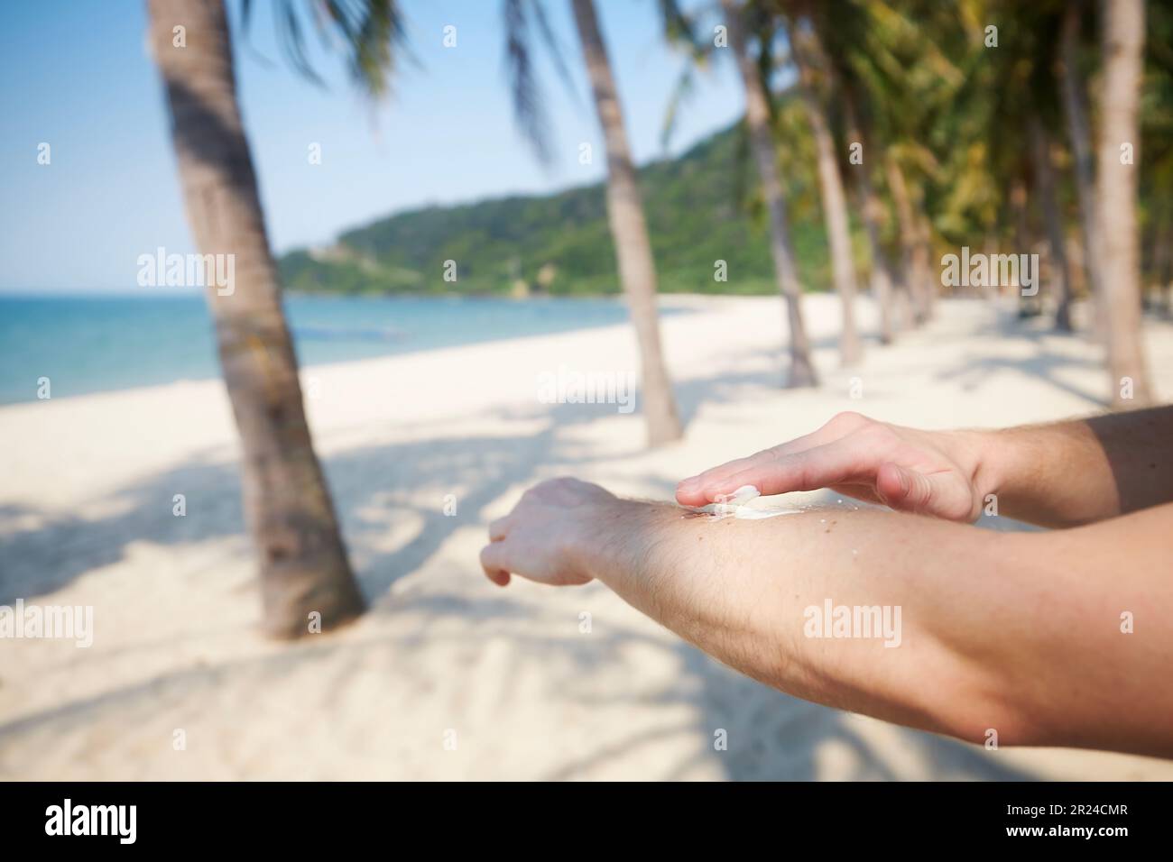 Primo piano della mano durante l'applicazione della crema solare. Cura della pelle durante le vacanze estive sulla bella spiaggia di sabbia con palme in una destinazione tropicale. Foto Stock