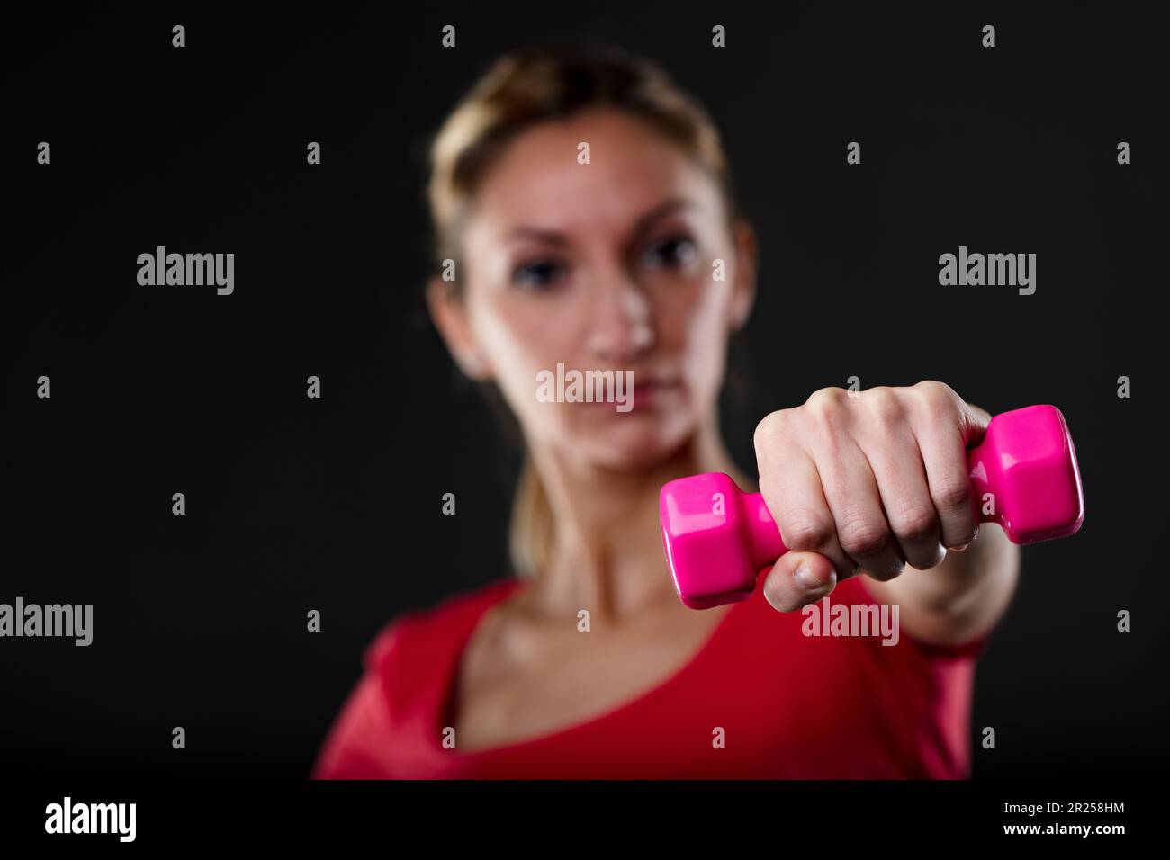 Donna sollevamento manubri rosa, sfondo sfocato. Bionda, parte superiore rossa, flaunts stomaco piatto, fiero degli obiettivi di idoneità. Dopo Mens sana in corpore sano mo Foto Stock