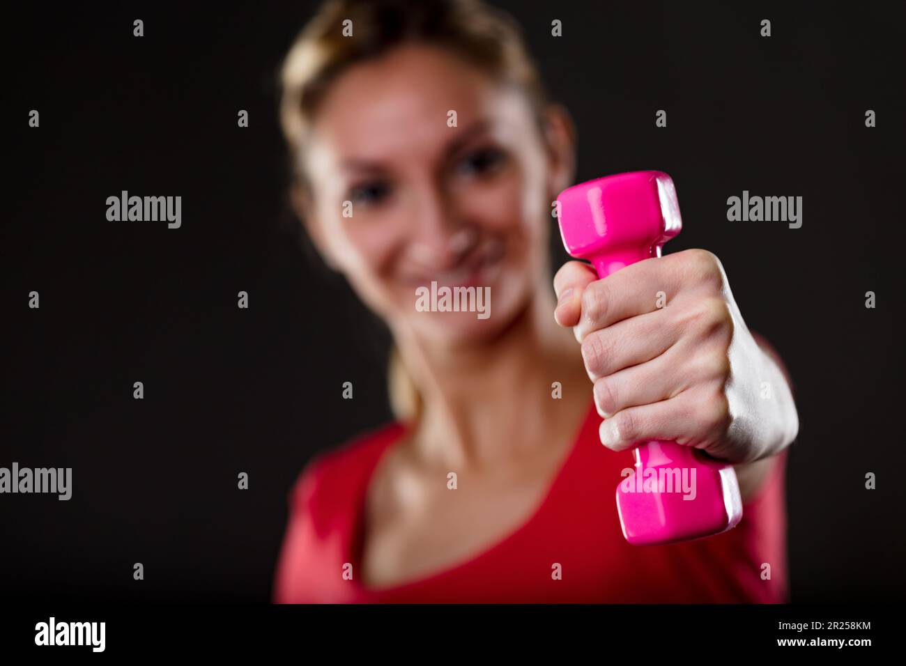 Donna sollevamento manubri rosa, sfondo sfocato. Bionda, parte superiore rossa, flaunts stomaco piatto, fiero degli obiettivi di idoneità. Dopo Mens sana in corpore sano mo Foto Stock
