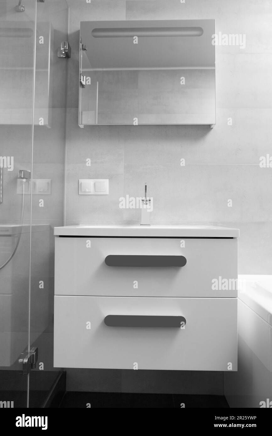 Parte dell'interno di un bagno moderno, un lavabo bianco con armadio e cassetti, un rubinetto cromato, un grande specchio illuminato e una doccia in vetro Foto Stock