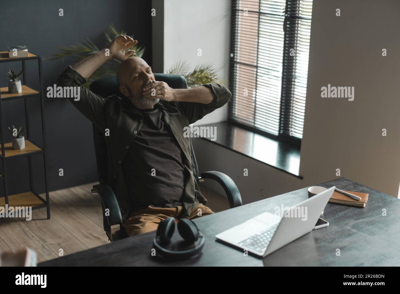 Uomo anziano stanco con la barba d'argento sbadigliano mentre lavora a distanza dalla comodità di casa. Realtà di fatica ed esaurimento che possono accompagnare il lavoro remoto. . Foto di alta qualità Foto Stock