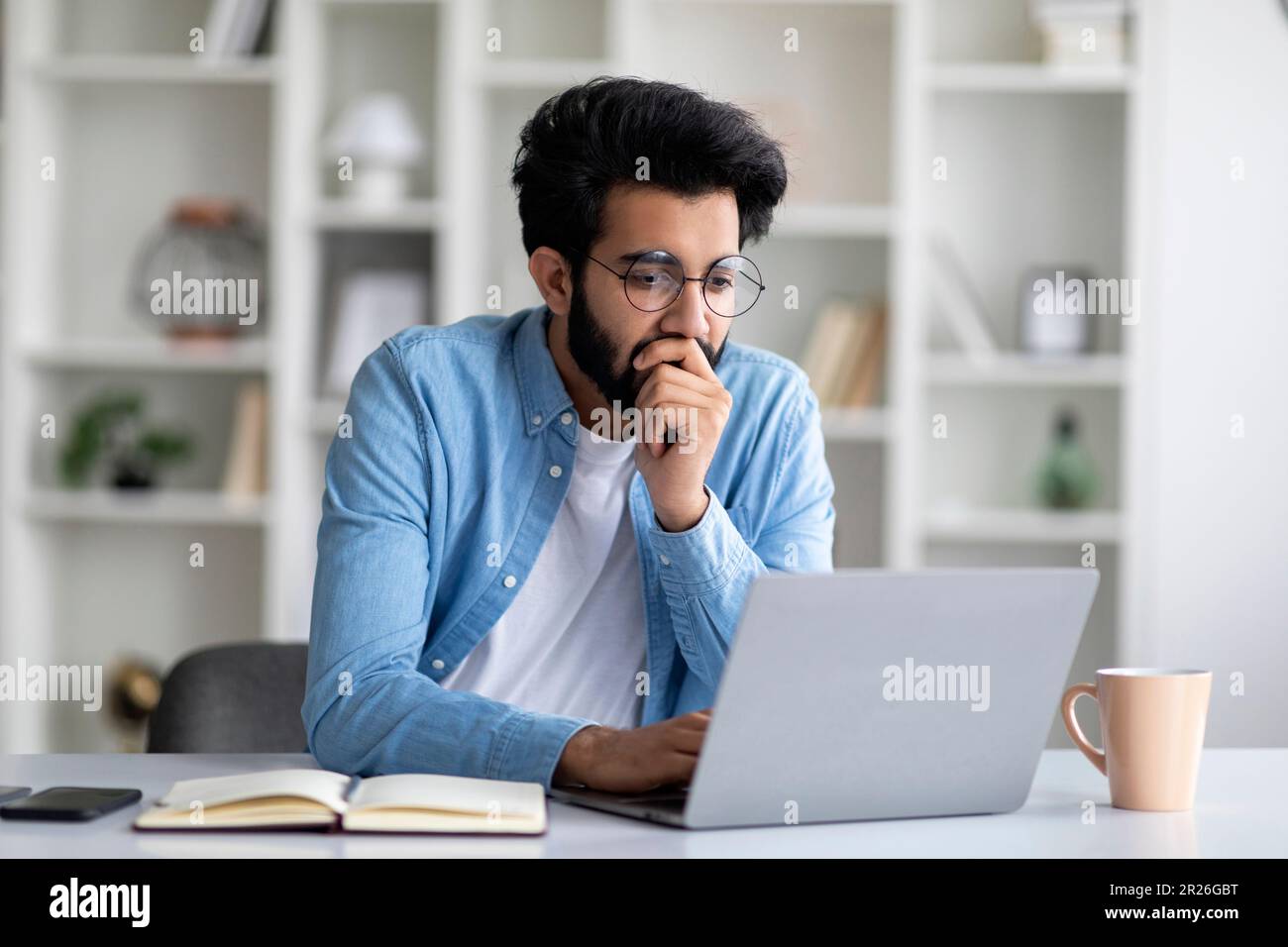 Ritratto di un giovane uomo indiano focalizzato in occhiali che lavorano su laptop Foto Stock