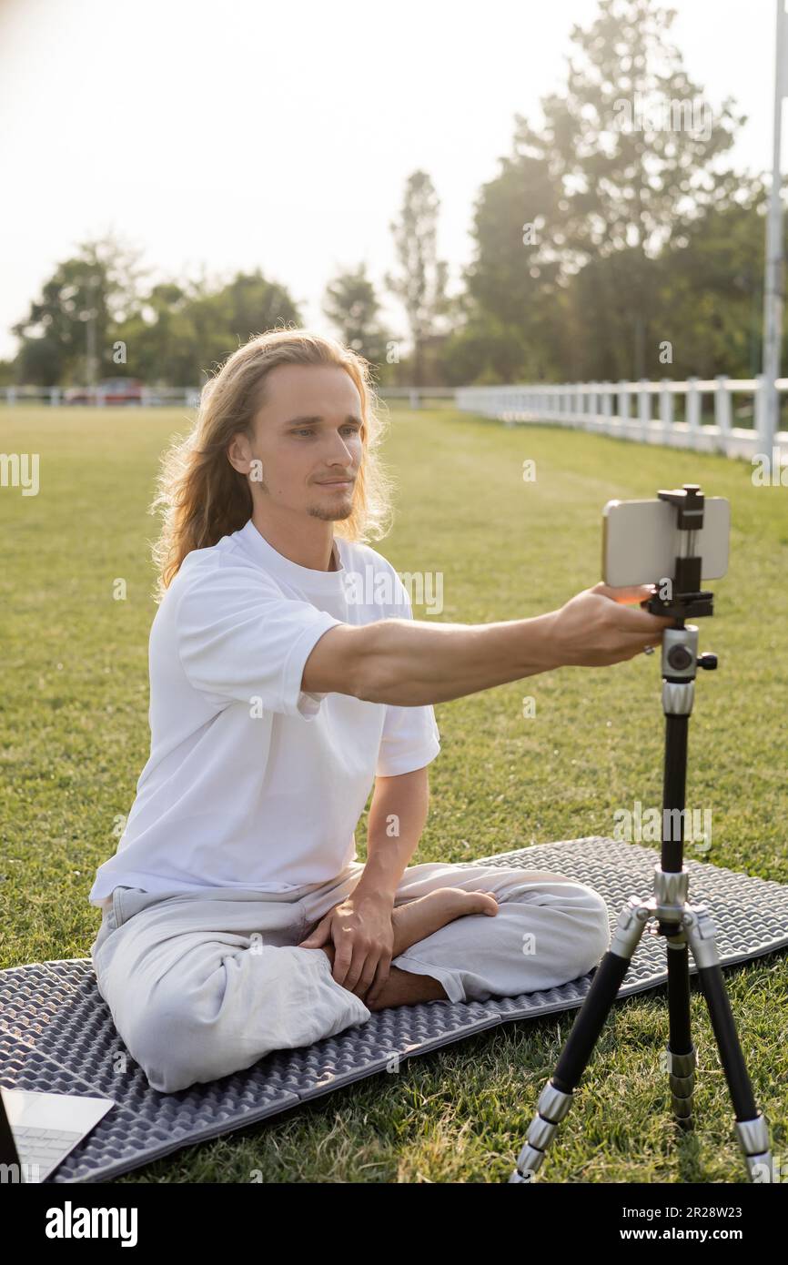 yoga vlogger seduto in posa facile e regolare treppiede con smartphone, mentre seduto su erba verde all'aperto Foto Stock