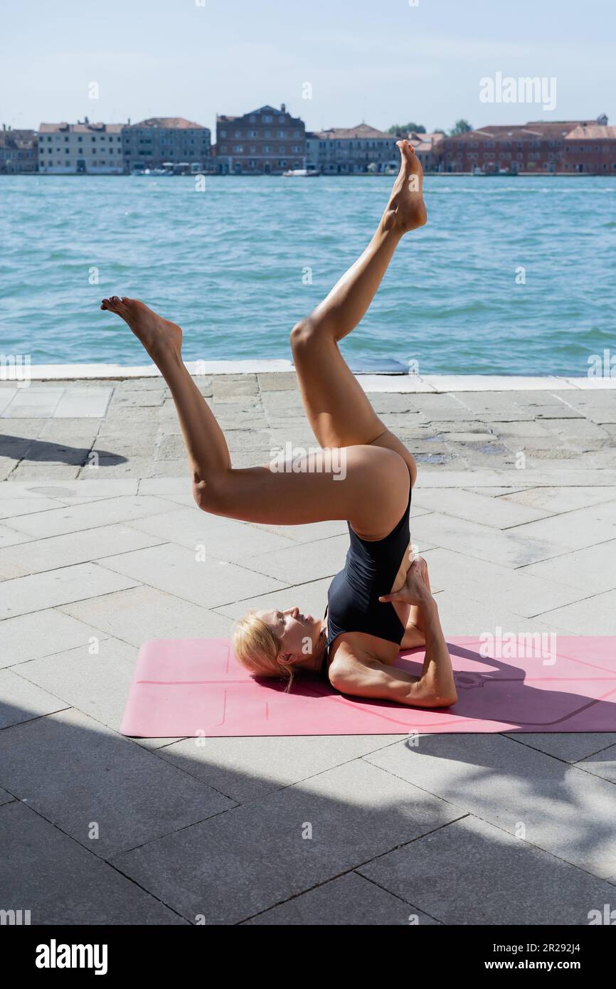 Donna bionda in bodysuit facendo yoga asana su tappetino rosa a Venezia Foto Stock