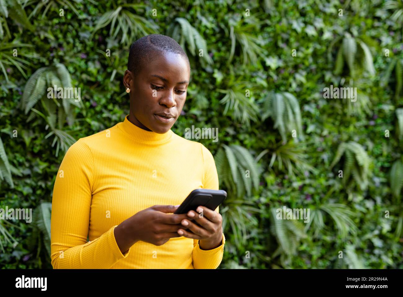 Donna d'affari afroamericana con i capelli corti che testano sullo smartphone mentre si levano in piedi contro le piante Foto Stock