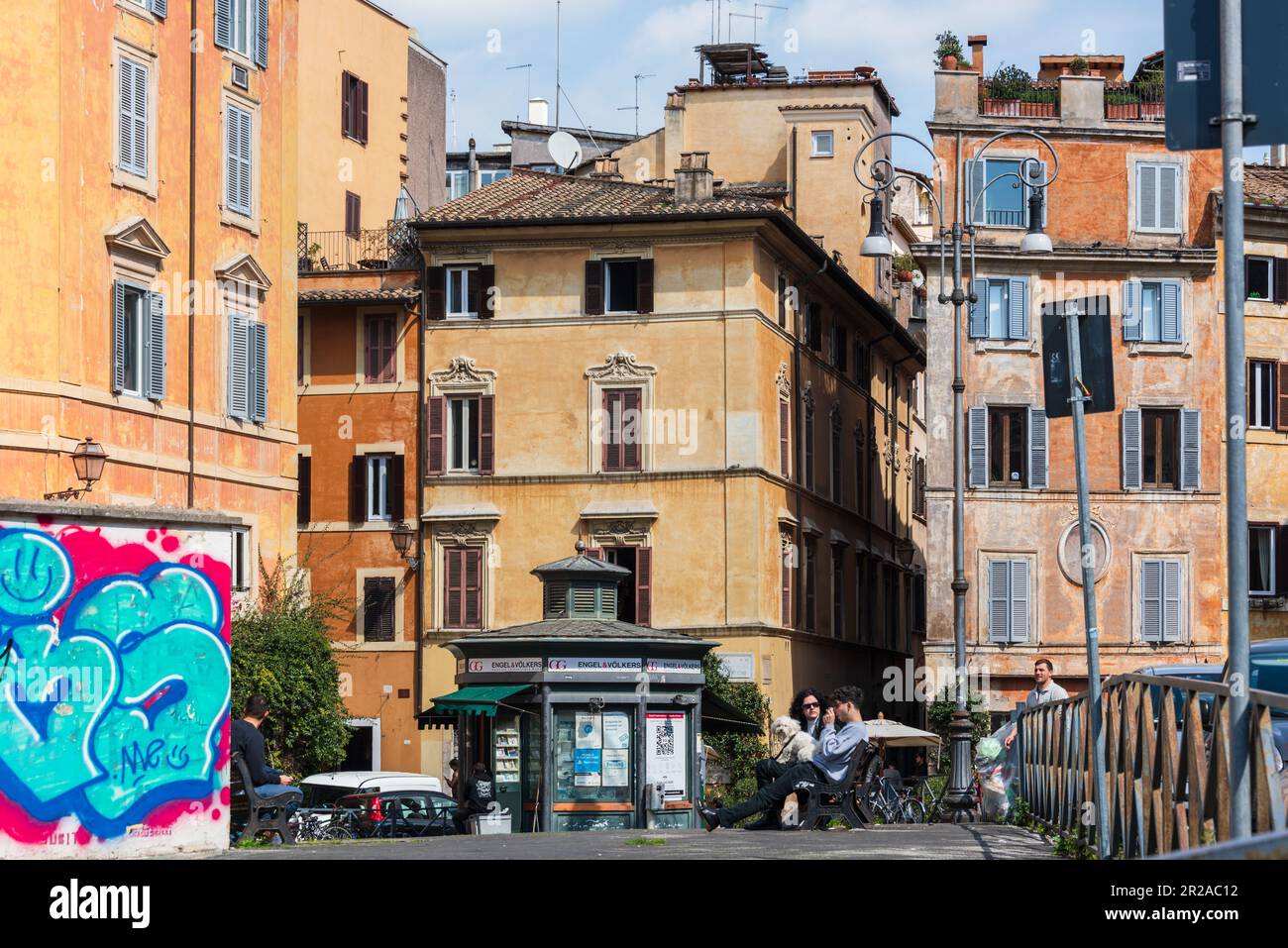 ROM, Italien, März 2023 Straßenszene ein Kiosk mit Gastronomie und Außensitzplätzen Ein Paar mit einem Hund auf einer Bank Sitzend, Huuswand mit Groß Foto Stock