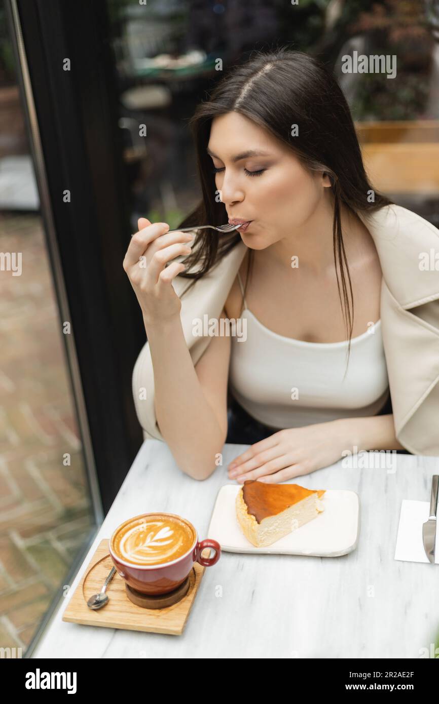 giovane donna con i capelli lunghi che mangiano cheesecake vicino alla tazza di cappuccino sul tavolo del bistro mentre si siede nella giacca di pelle accanto alla finestra all'interno del moderno Foto Stock