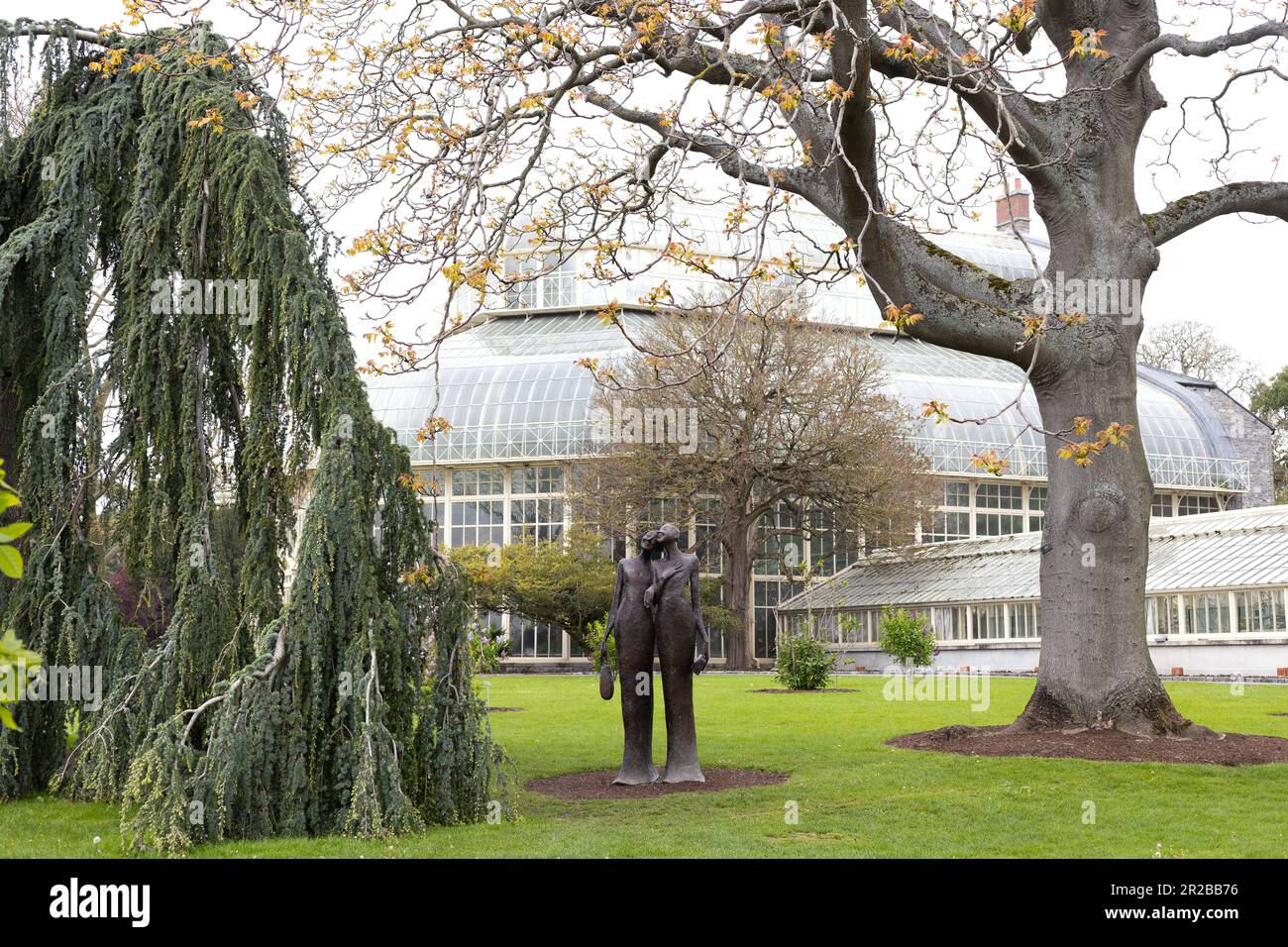 Una statua e alberi di fronte a una serra presso i Giardini Botanici nazionali di Dublino, Irlanda. Foto Stock