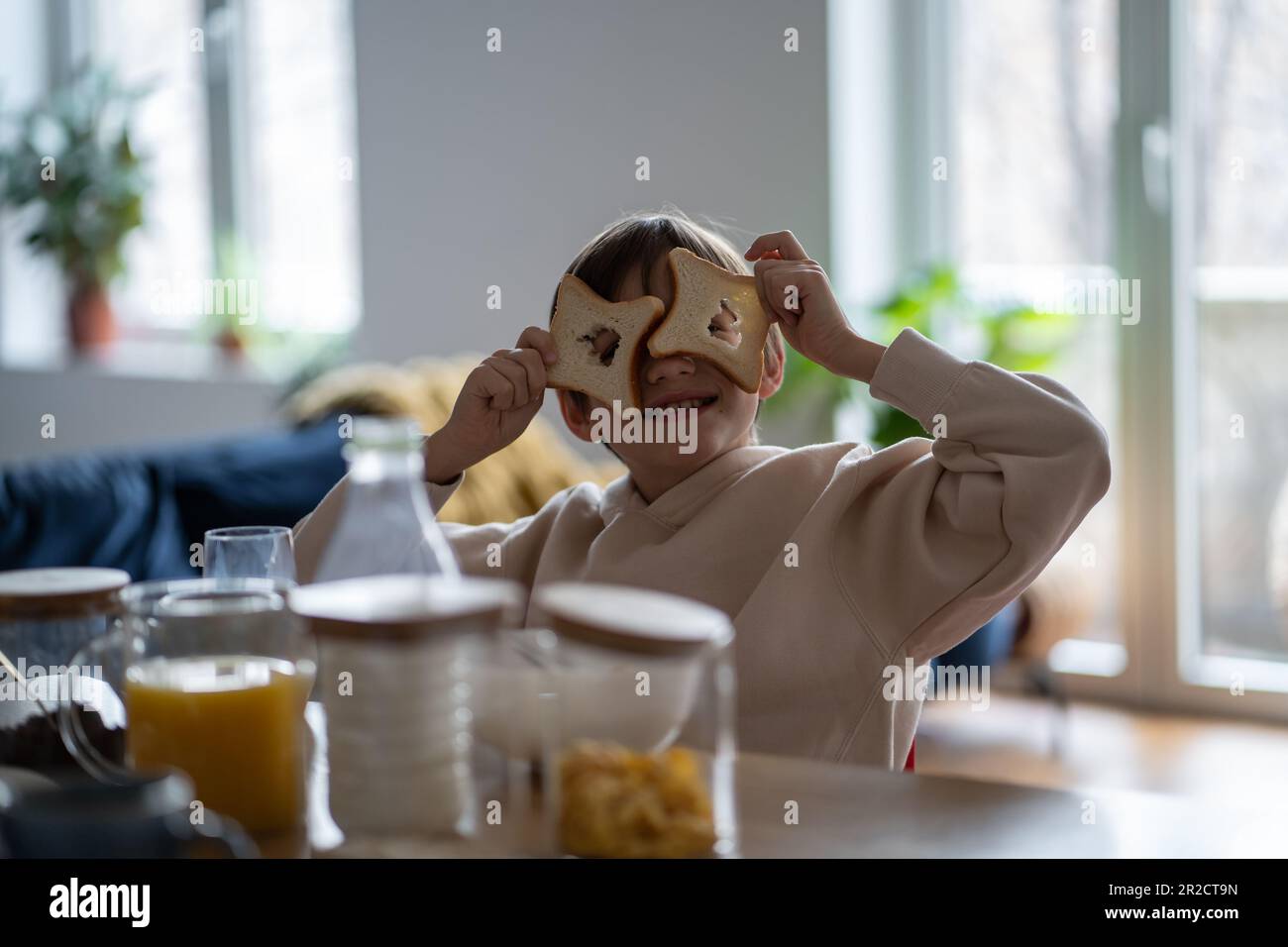 Figlio ragazzo imbrogliando giocando con pezzi di pane che guardano attraverso i fori della macchina fotografica seduto al tavolo. Foto Stock