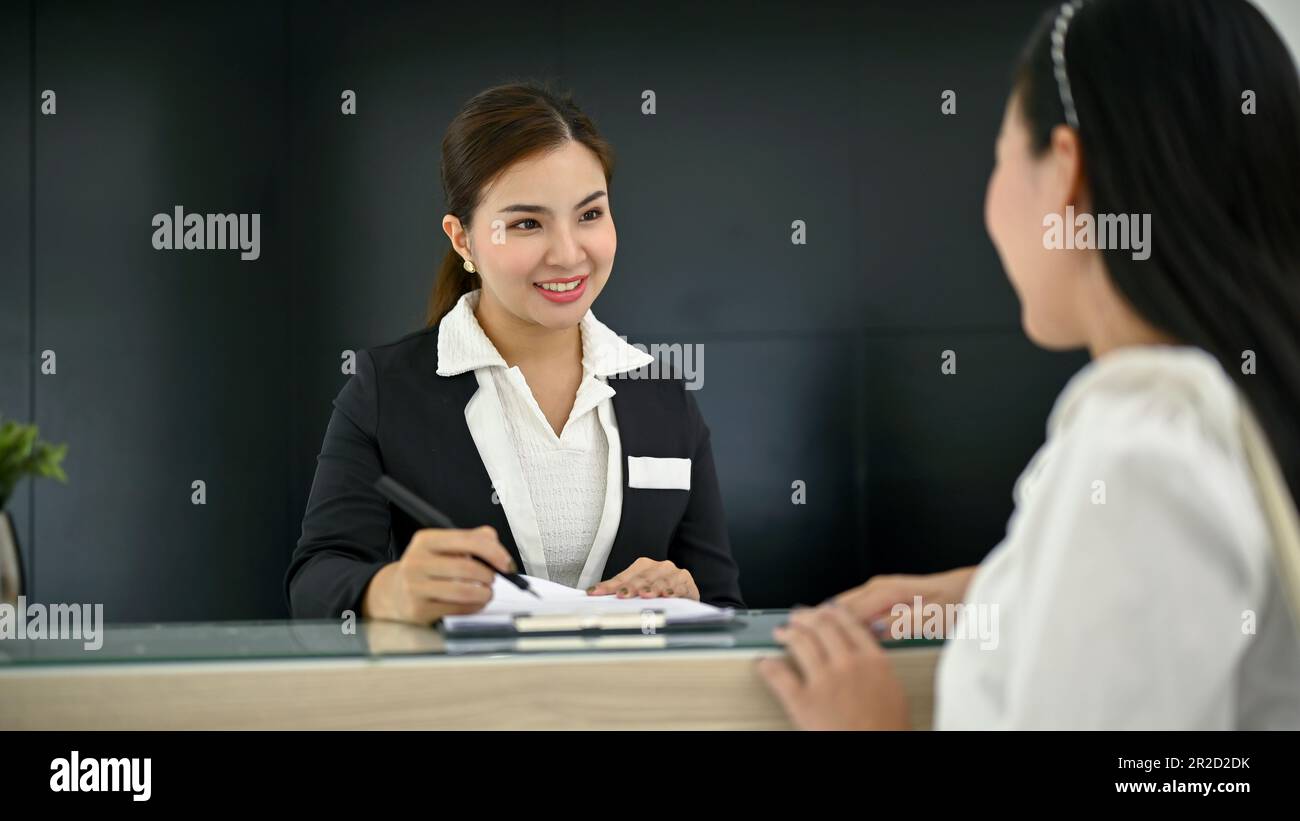 Una bella receptionist femminile asiatica millenaria sta parlando e chiedendo informazioni a un cliente nella lobby. Foto Stock