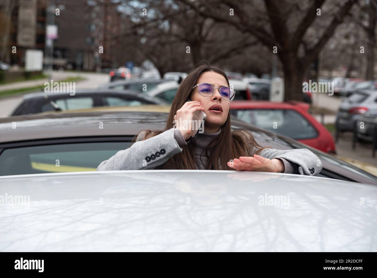 Giovane donna d'affari nervosa in piedi accanto alla sua auto dopo l'orario di lavoro che parla al telefono discutendo con i dipendenti perché deve tornare a lavorare Foto Stock