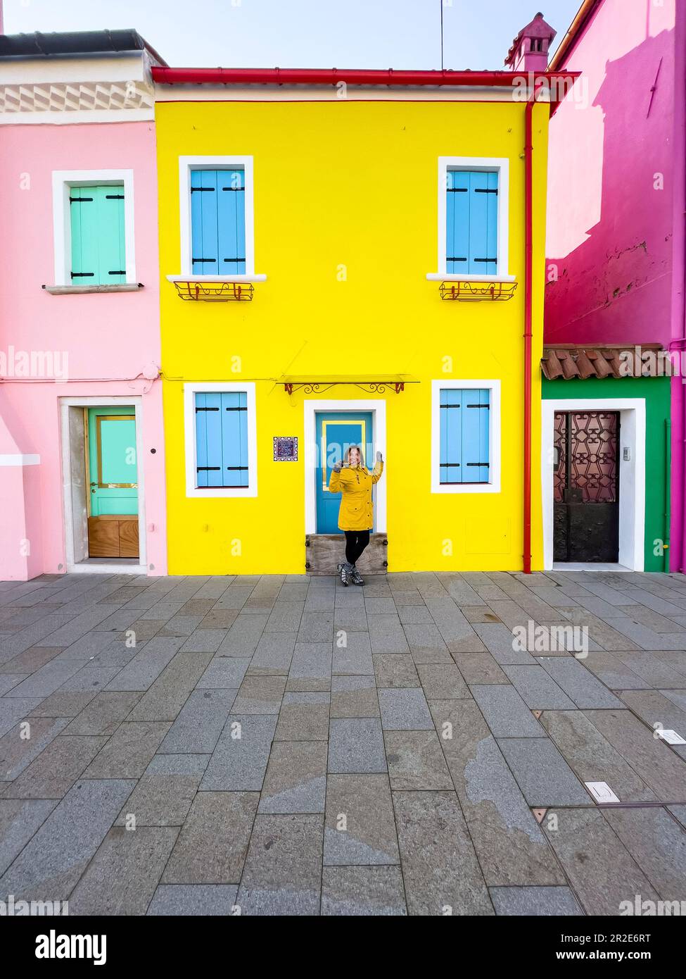 Donna sorridente che posa di fronte alla casa gialla a Burano Island. Felice viaggiatore turista femmina con impermeabile e occhiali da sole in posa e divertirsi Foto Stock