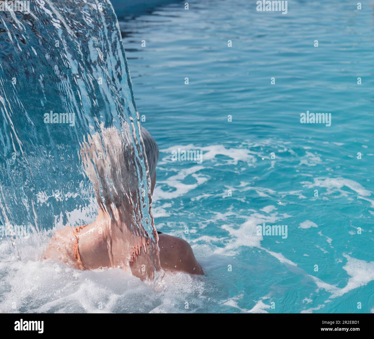 Donna anziana con i capelli grigi che godono il flusso di acqua che cade sulle sue spalle. Vista posteriore. Idromassaggio nella piscina termale esterna. Anziani attivi Foto Stock