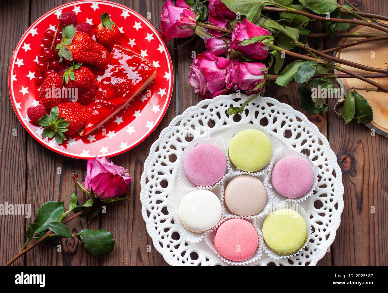 Fetta di torta di fragole, macaron e fiore di rosa su piatto rosso posto sul tavolo di legno dall'alto. La colazione era bellissima. Stile piatto. Foto Stock