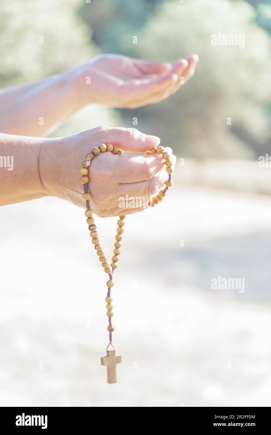 Mani di donna che tengono un rosario cristiano con una croce, pregando in un campo al crepuscolo Foto Stock