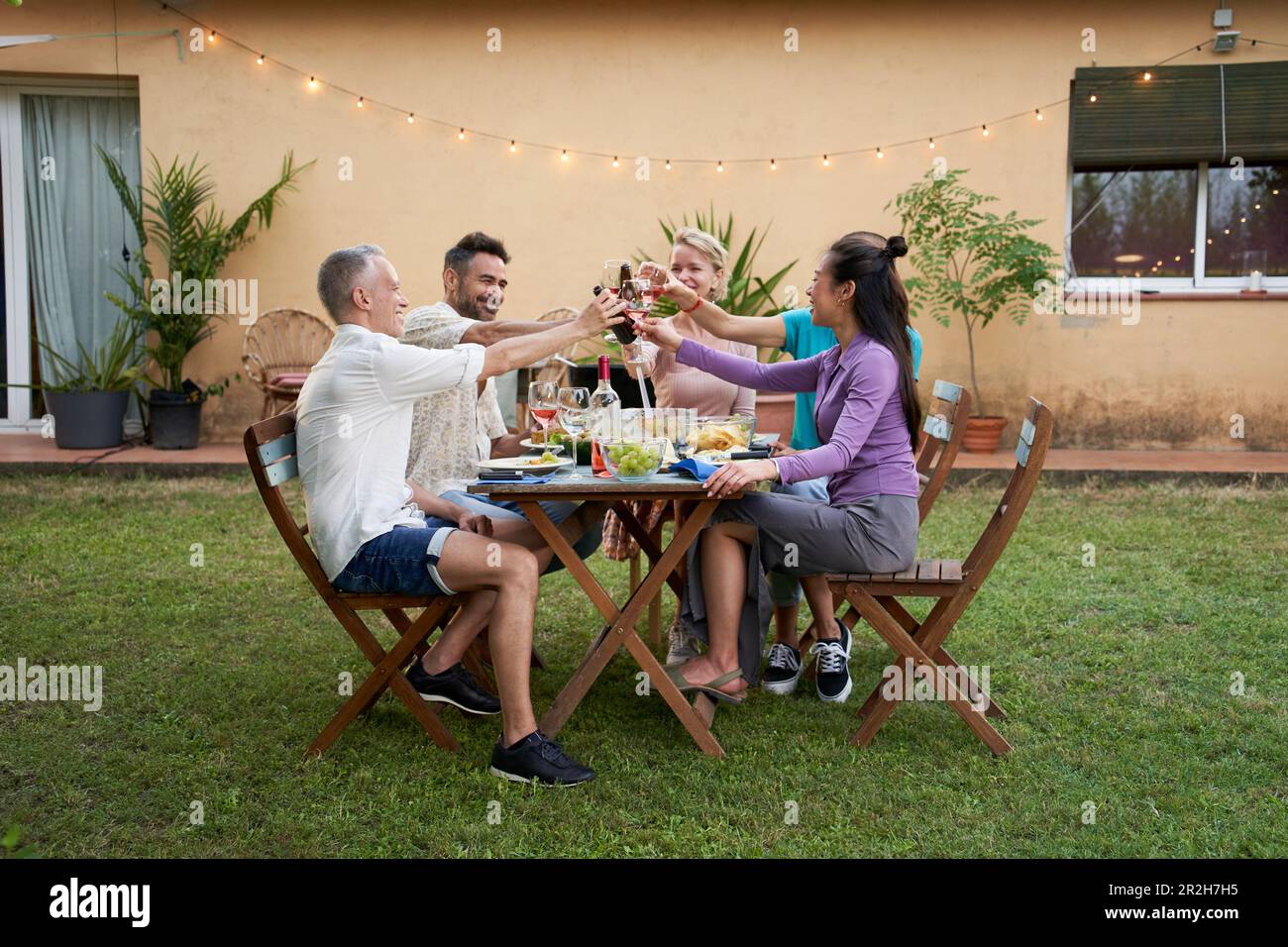 Un gruppo di amici di mezza età brinda con il vino raccolto intorno a un tavolo da giardino in una serata estiva per condividere un pasto e avere un buon tempo insieme. Foto Stock