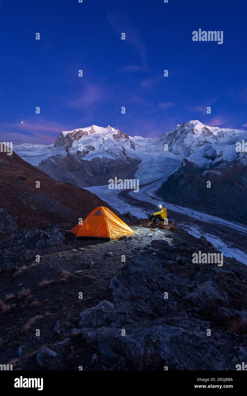 Escursionista con lampada frontale seduto accanto alla tenda di fronte al massiccio del Monte Rosa, al ghiacciaio Gorner (Gornergletscher) e alle cime del Lyskamm al crepuscolo Foto Stock