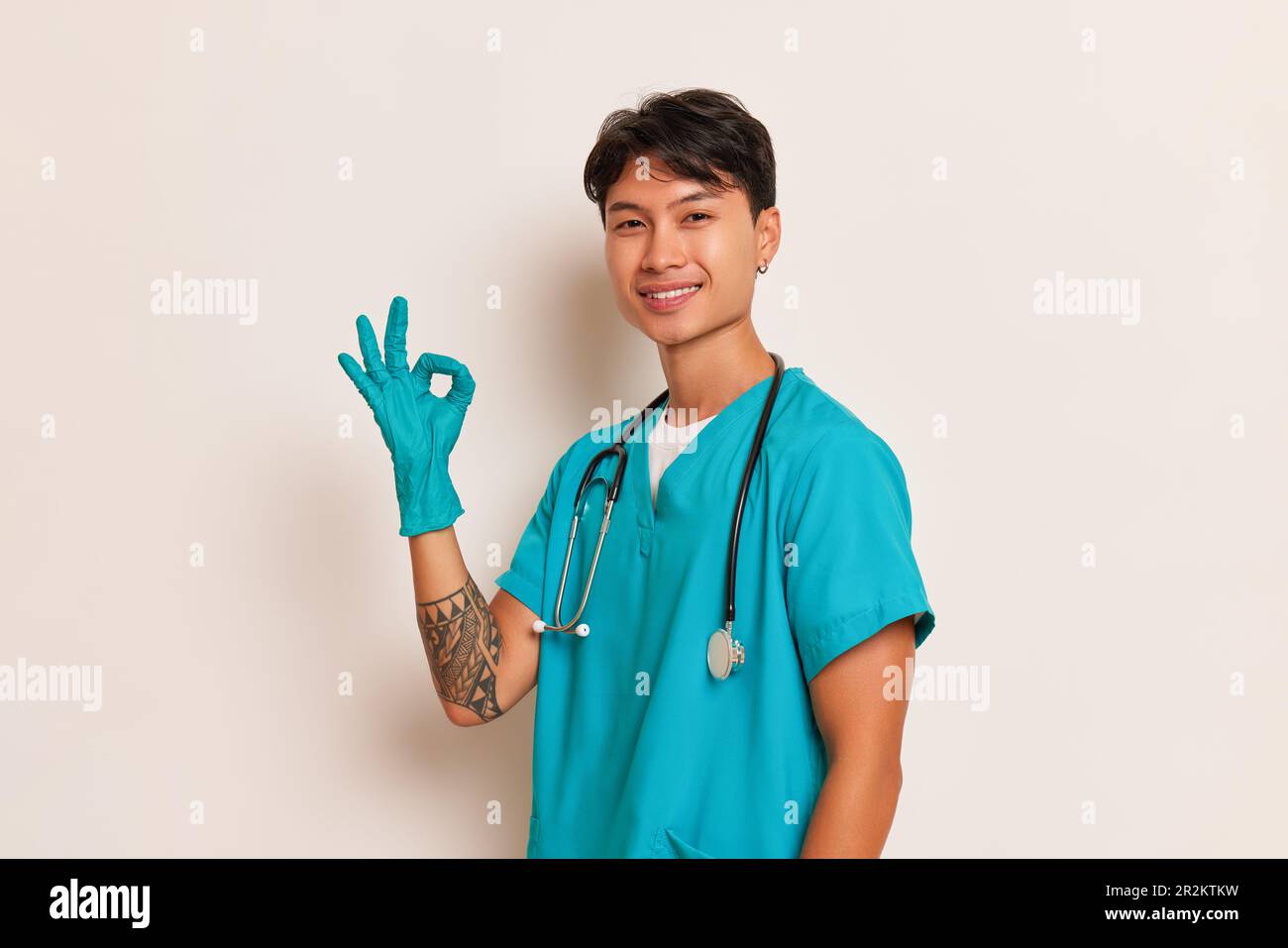 Giovane operatore medico con stetoscopio sul collo e guanti in lattice sulle mani posa a parete bianca e mostra ok segno con una mano, professionale Foto Stock