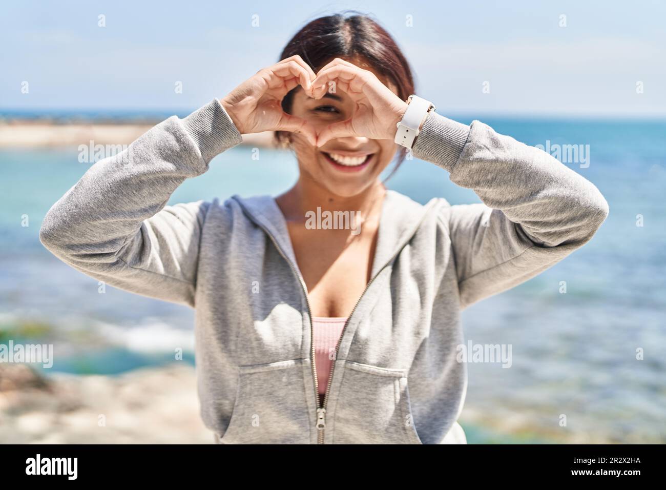 Giovane donna ispanica che indossa abbigliamento sportivo che fa simbolo del cuore con le mani al mare Foto stock Alamy