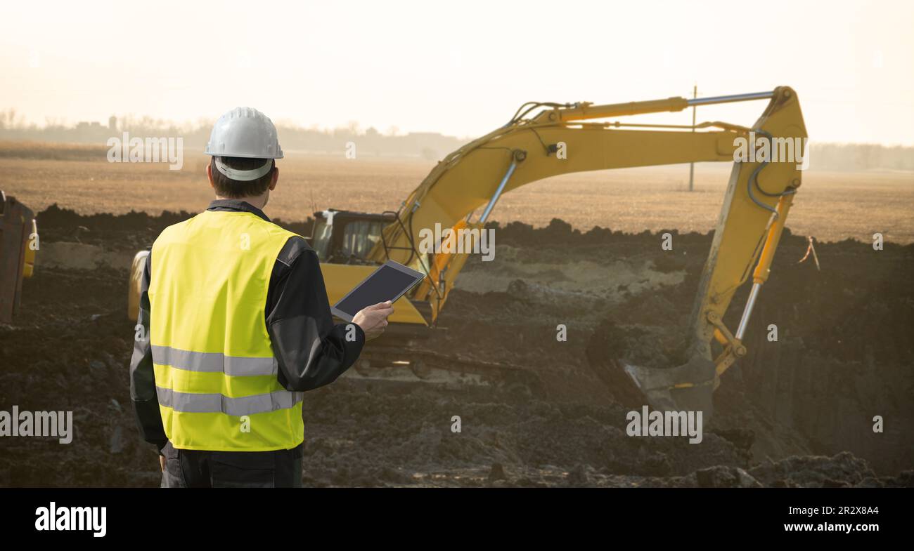 Ingegnere in un casco con un tablet digitale accanto all'escavatore da cantiere. Foto di alta qualità Foto Stock