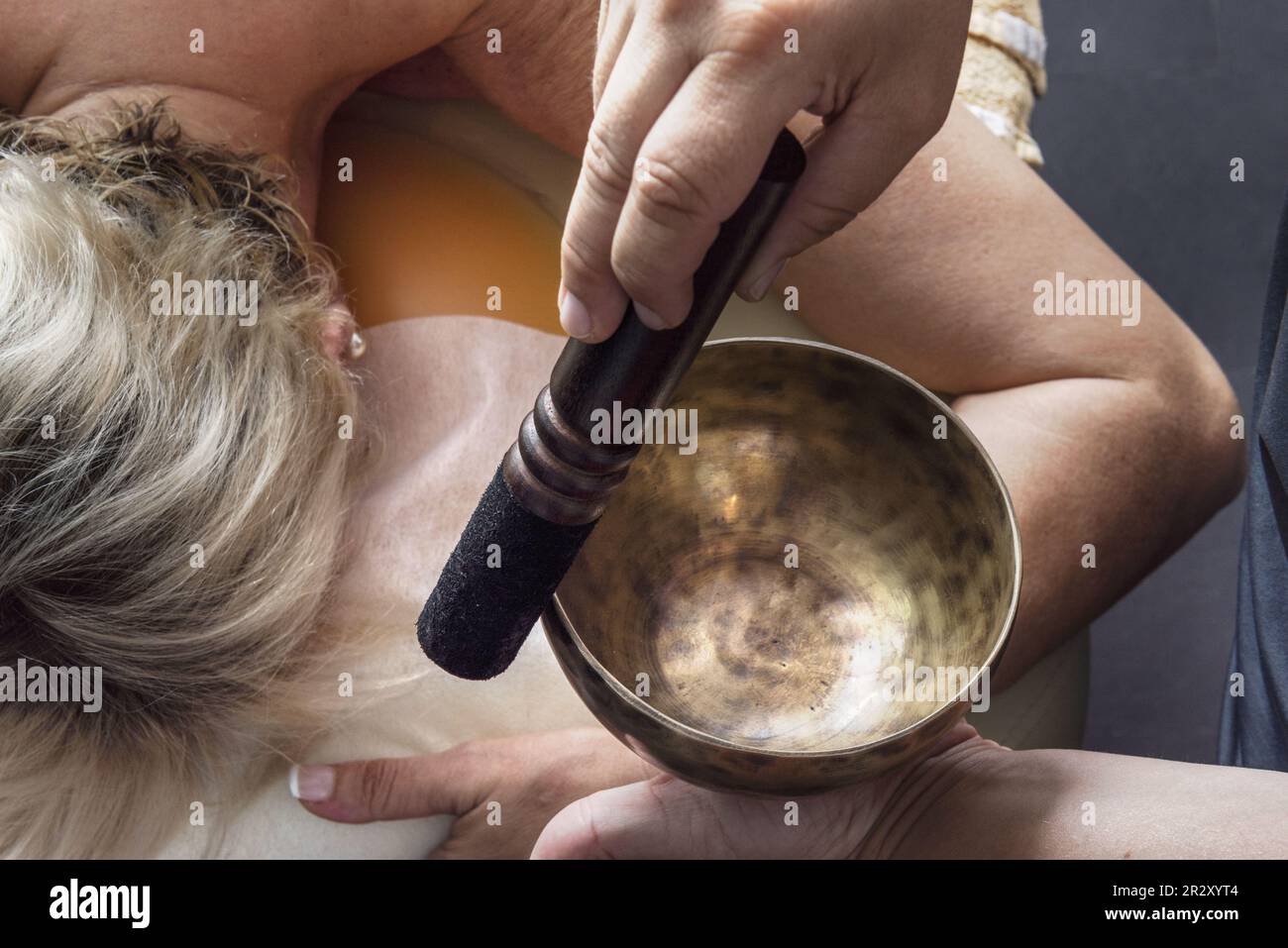 Una massaggiatrice che esercita una pressione su una ciotola cantante sul lato della testa di un paziente sdraiato su un tavolo Foto Stock