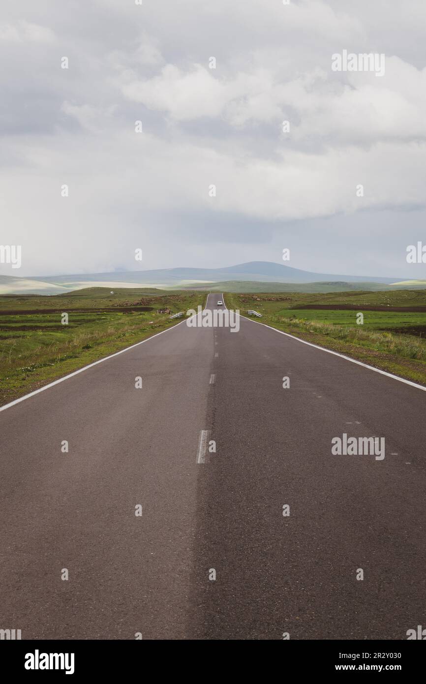 Strada asfaltata dritta attraverso verdi praterie dell'altopiano di Javakheti con auto bianca in lontananza e le montagne del Caucaso, Georgia. Foto Stock