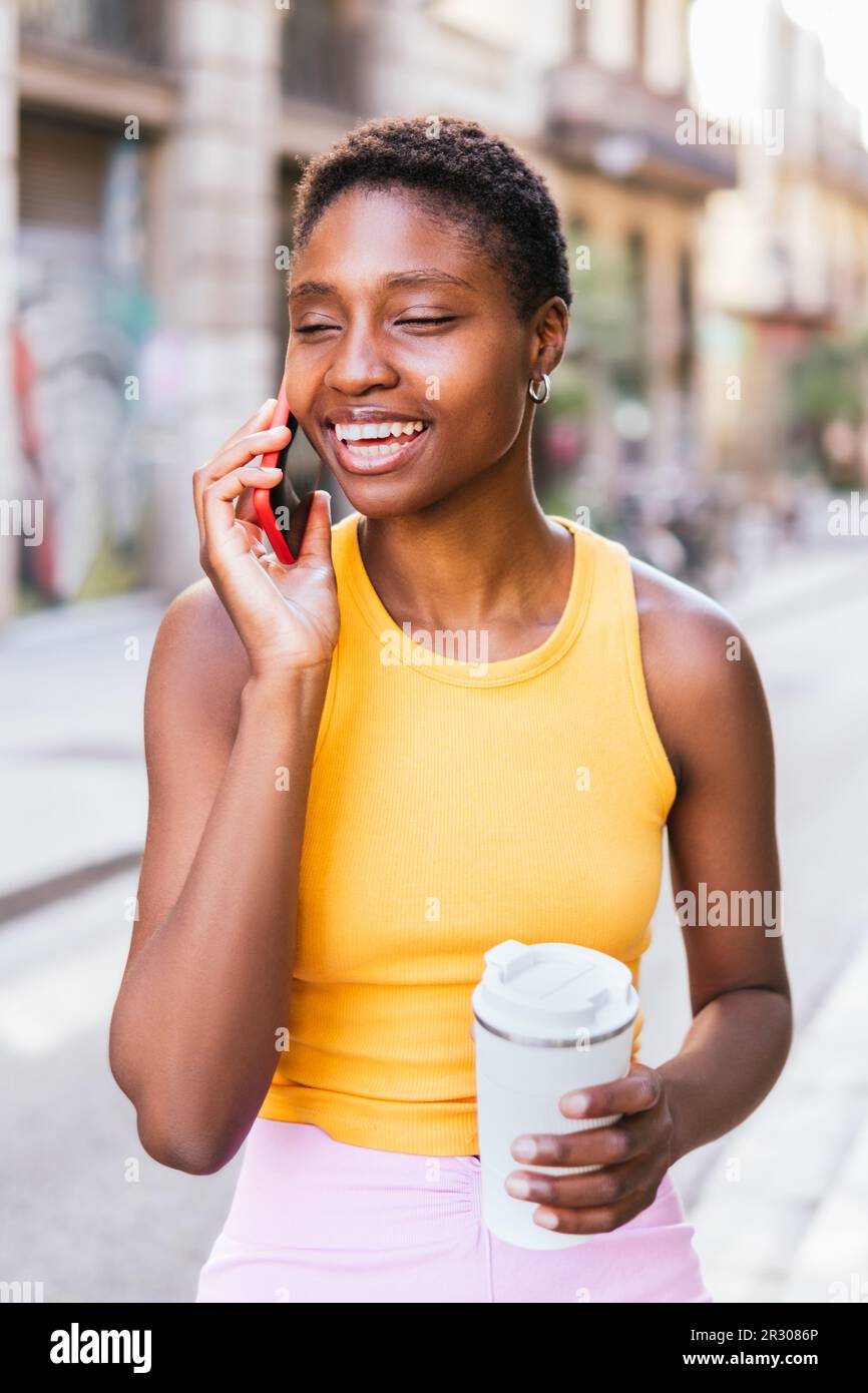 Pendolari afro-americani con tazza di caffè riutilizzabile per strada. Sta facendo una conversazione telefonica e sorridendo Foto Stock