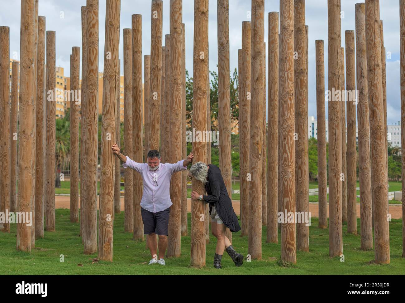 Una coppia di mezza età gioca tra pali decorativi in legno in un giardino. Appoggia le mani sui pali, abbraccia un palo. Foto Stock