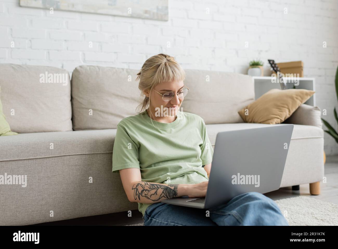 donna tatuata con capelli biondi e corti, scopa e occhiali a scrivere sul  computer portatile mentre si siede sul tappeto vicino a un comodo divano  nel moderno soggiorno Foto stock - Alamy