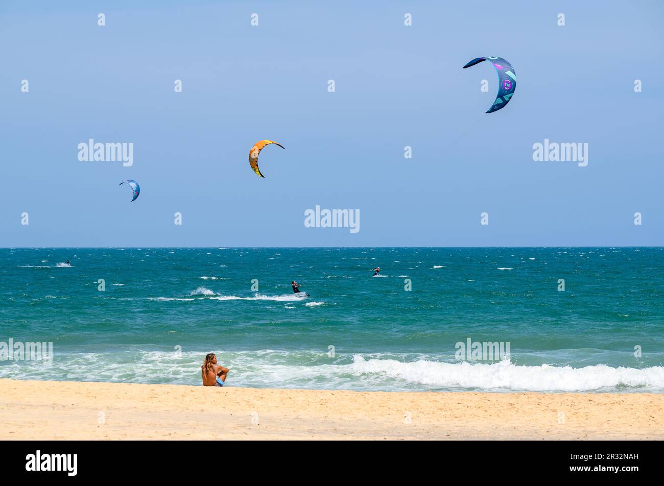 Un uomo locale si siede da solo sulla spiaggia osservando i kitesurfers in azione in mare in Mui NE, Vietnam. Foto Stock