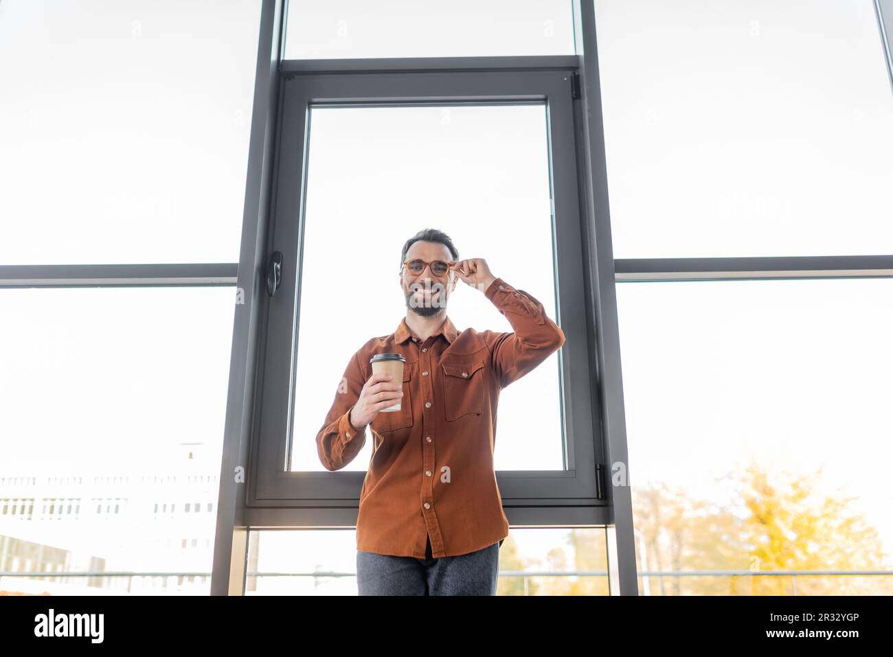 vista dal basso angolo di un allegro uomo d'affari in camicia alla moda in piedi con caffè per andare vicino a grandi finestre da ufficio, regolazione degli occhiali e Foto Stock