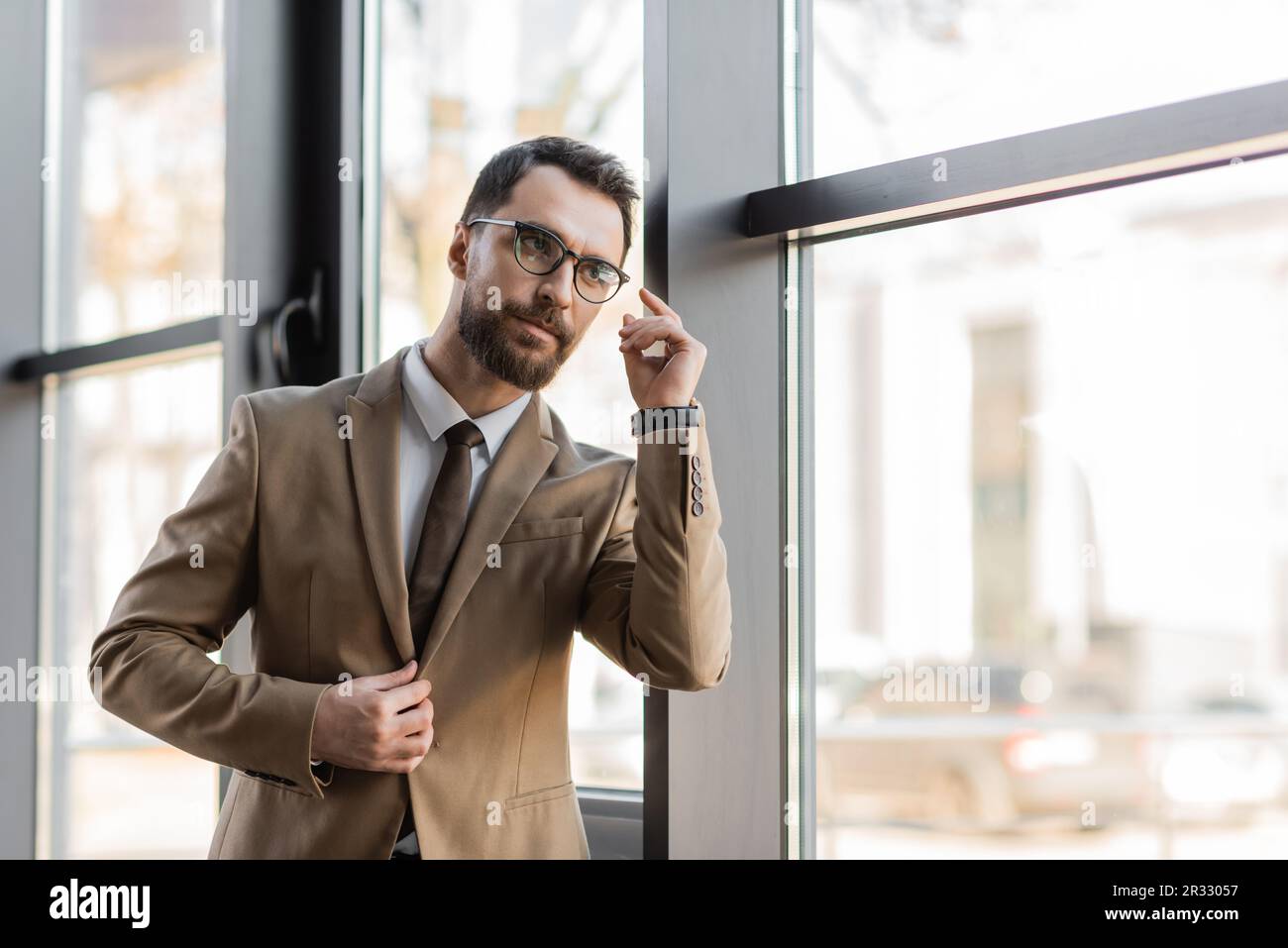 imprenditore premuroso e bearded in abbigliamento di affari alla moda come blazer beige, cravatta e occhiali che guardano lontano vicino a grandi finestre in moderno Foto Stock