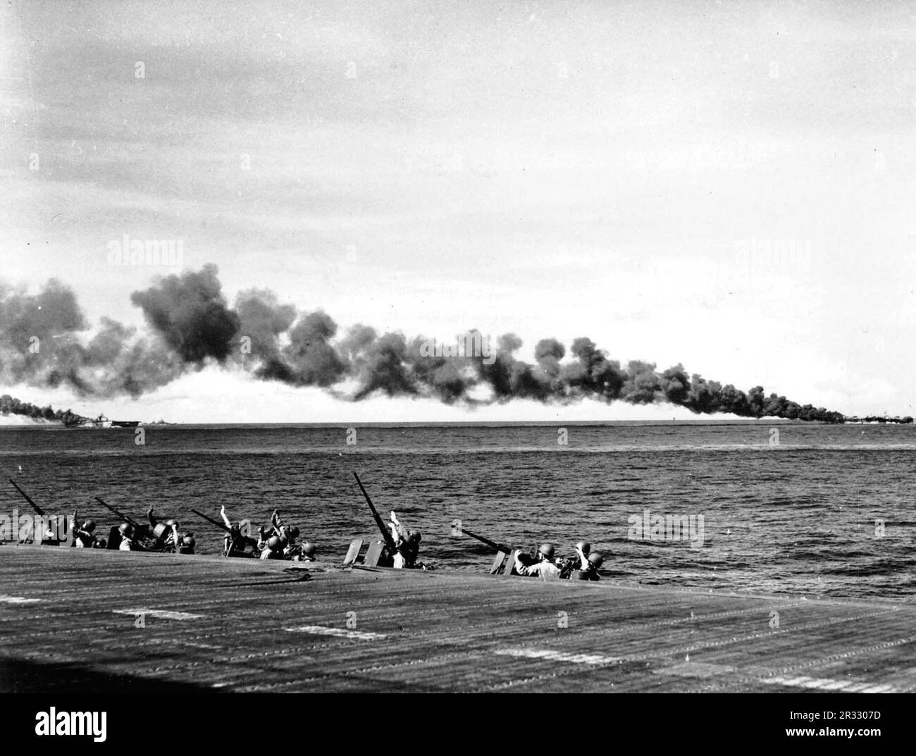 Membri del distaccamento Marino a bordo degli Stati Uniti La portaerei Navy USS Enterprise (CV-6) protegge gli occhi dal sole mentre scannerizzano i cieli per cercare velivoli kamikaze che si tuffano fuori dal sole. Si noti che i vettori USS Franklin (CV-13), a sinistra, e USS Belleau Wood (CVL-24) bruciano dopo essere stati colpiti da aerei kamikaze. Foto Stock