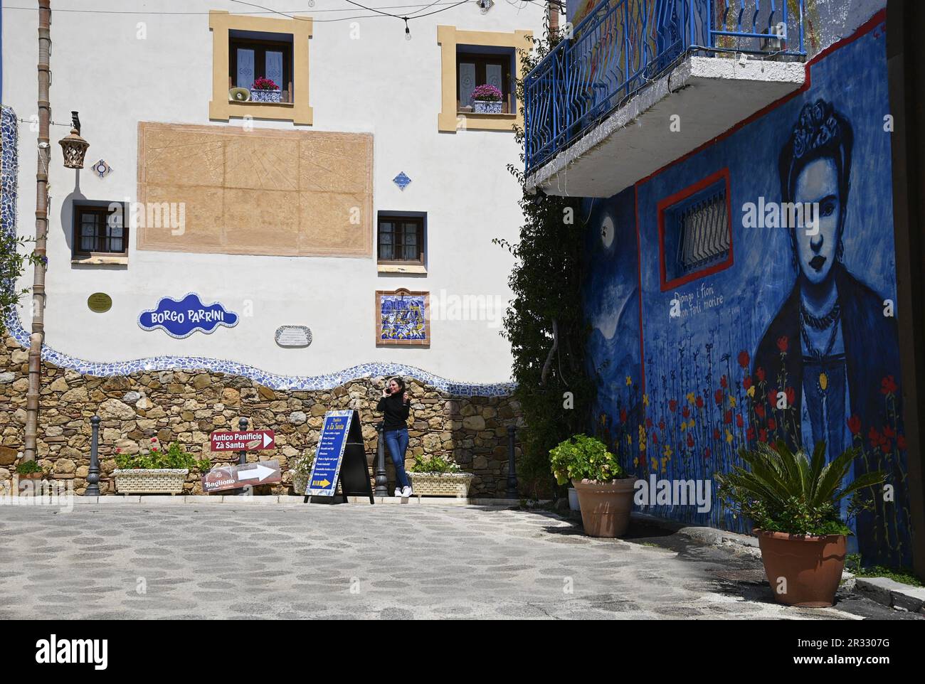 Giovane ragazza che parla sul suo cellulare sulla piazza principale con il famoso murale Frida Kahlo a Borgo dei Parrini in Sicilia. Foto Stock