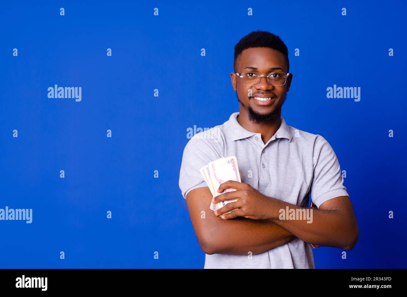 Giovane bell'uomo africano sorridente mentre tiene contanti con le braccia incrociate, in piedi su uno sfondo isolato blu Foto Stock