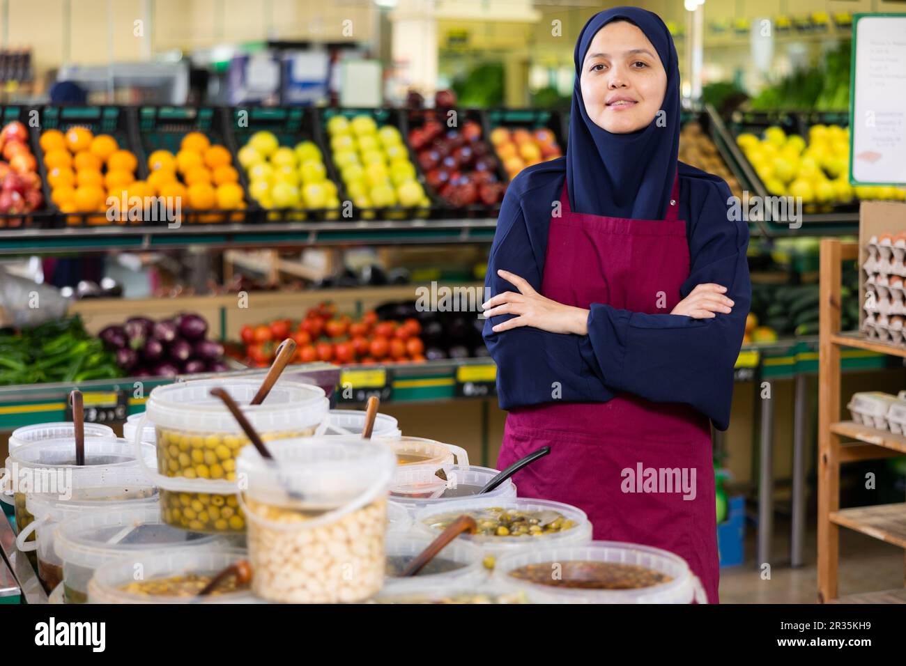 Nel negozio di verdure accanto alle olive in salamoia per peso si trova donna venditore in hijab e sorride affettuosamente Foto Stock