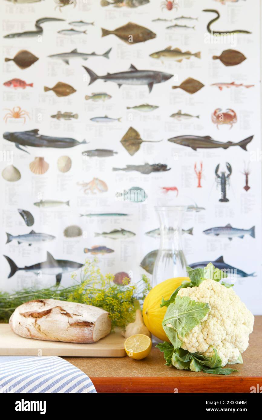 Pane, cavolfiore, aneto, melone, limone e una caraffa d'acqua di fronte alla carta da parati di pesce Foto Stock