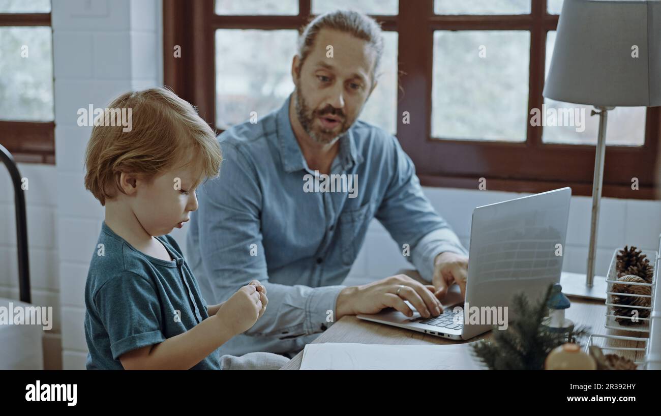 Il padre che lavora da casa utilizzando un computer portatile siede al tavolo con un simpatico piccolo figlio che gioca quasi a disturbare. Foto Stock