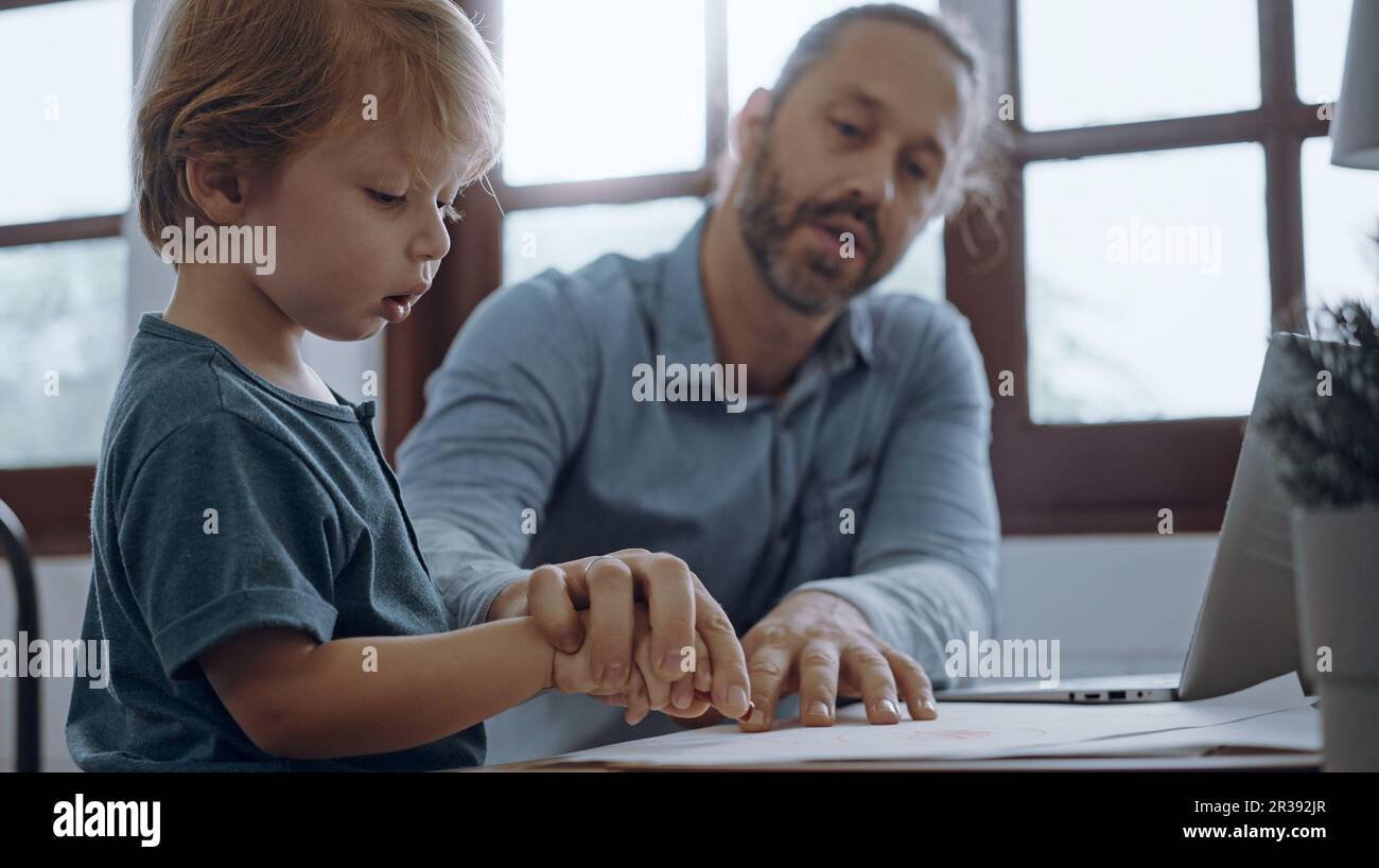 Il padre che lavora da casa utilizzando un computer portatile siede al tavolo con un simpatico piccolo figlio che gioca quasi a disturbare. Foto Stock