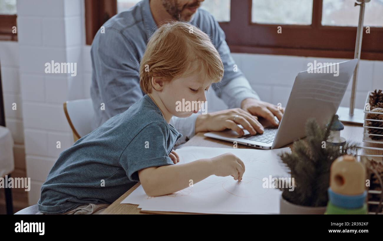 Il padre che lavora da casa utilizzando un computer portatile siede al tavolo con un simpatico piccolo figlio che gioca quasi a disturbare. Foto Stock