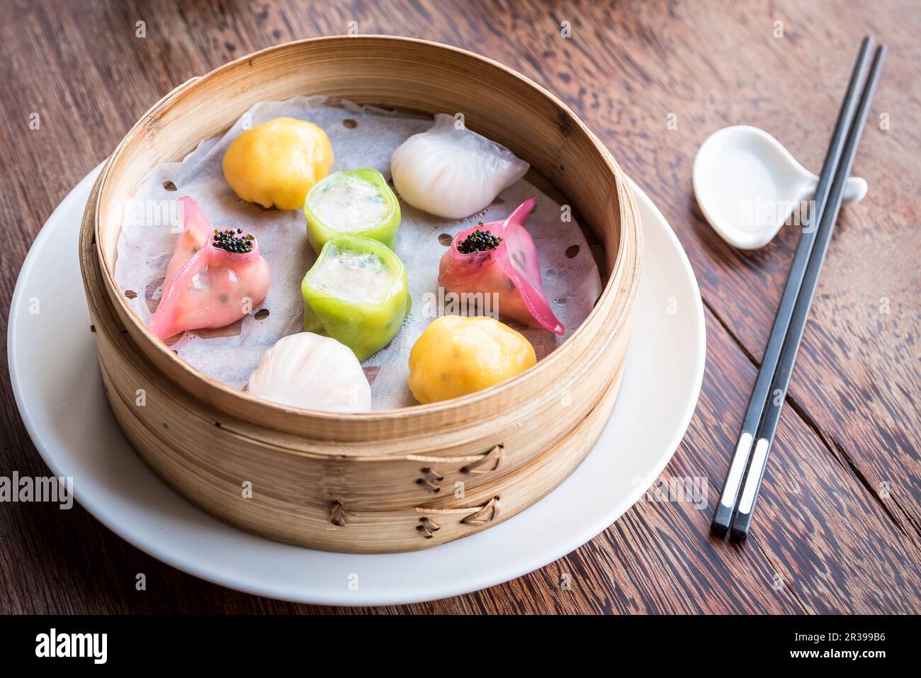 Gnocchi di dim sum assortiti e colorati in una vaporiera di bambù su un tavolo di legno con bacchette Foto Stock