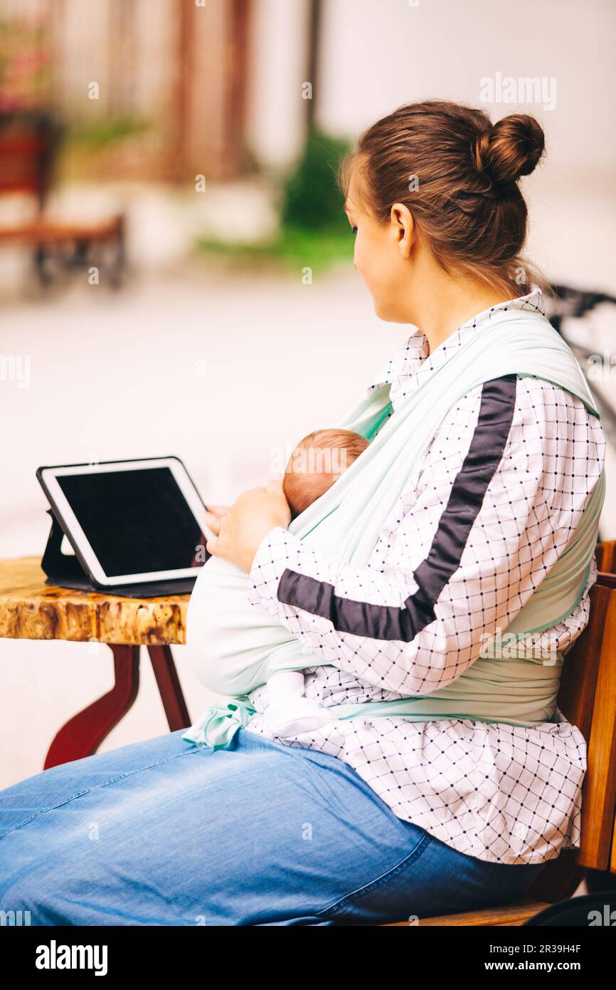 Giovane madre con bambino piccolo in sling seduto al bar all'aperto e di lavoro utilizzando un tablet Foto Stock