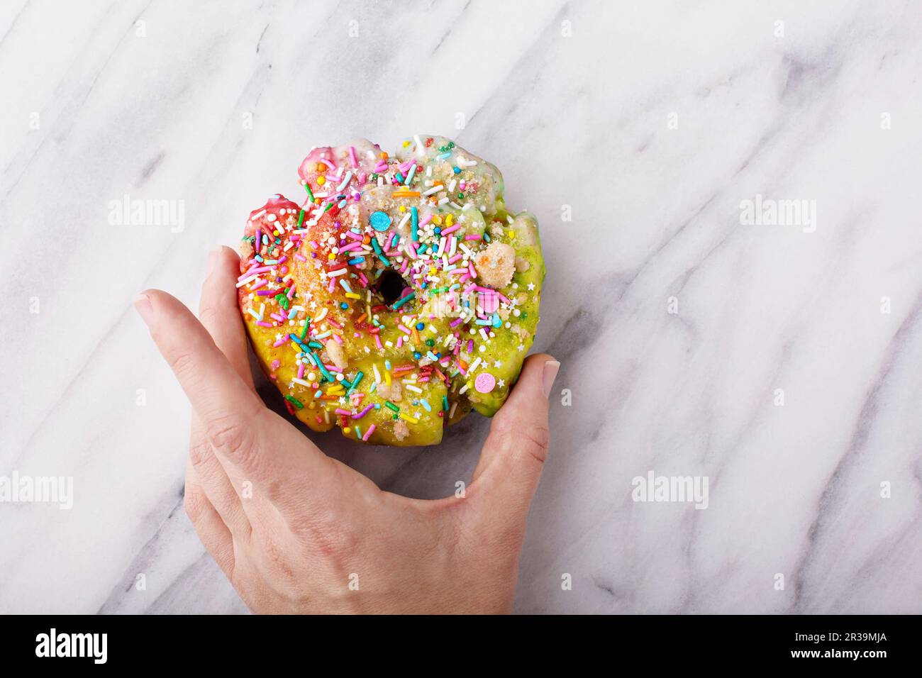 Donut di unicorno colorato e festoso con spruzzi sulla superficie di marmo con la mano di una donna che lo tiene, tendenza alimentare unicorno Foto Stock
