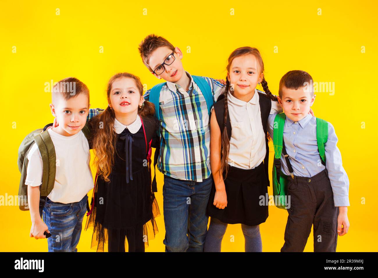 Gruppo di bambini felici in uniforme con borse da scuola abbracciando e guardando alla fotocamera su sfondo giallo Foto Stock