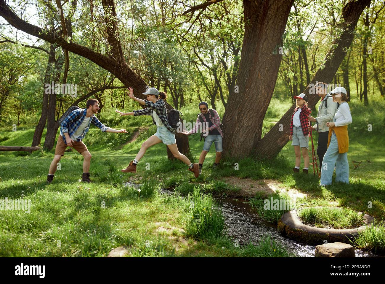 Giovani, uomini e donne, amici che fanno escursioni, camminano nella foresta, saltano sul fiume in una giornata calda Foto Stock