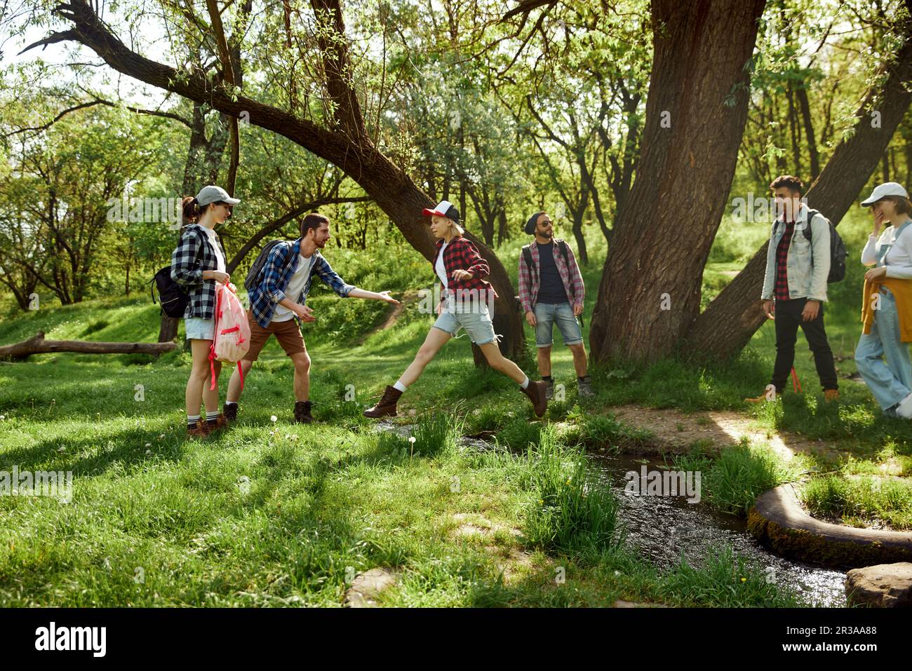 Giovani, uomini e donne, amici che fanno escursioni, camminano nella foresta, saltano sul fiume in una giornata calda Foto Stock
