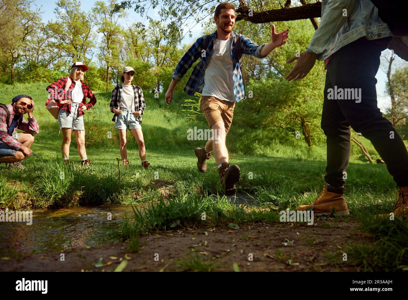 Gruppo di amici, giovani attivi che fanno escursioni, camminano nella foresta, saltano sul fiume e divertirsi all'aperto nella calda giornata primaverile Foto Stock