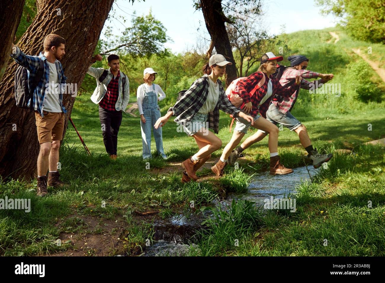 Gruppo di amici, giovani attivi in abiti casual andare a piedi, camminare nella foresta, saltare sul fiume in caldo giorno di primavera Foto Stock